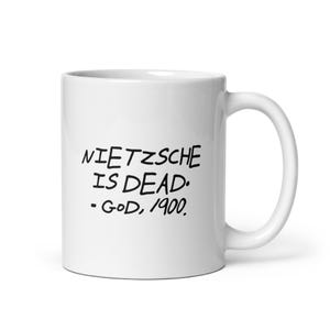Könnte beinhalten: Ein weißer Keramikbecher mit schwarzem, handschriftlichem Text, der NIETZSCHE IS DEAD. - GOD, 1900. lautet. Der Becher hat eine glatte, glänzende Oberfläche, einen zylindrischen Körper und einen geschwungenen Henkel auf der rechten Seite.