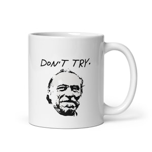 May include: A white ceramic mug featuring a black graphic illustration of a man's face with a beard and the text "DON'T TRY." printed in a simple, bold, black sans-serif font above the portrait. The mug has a curved handle on the right side.