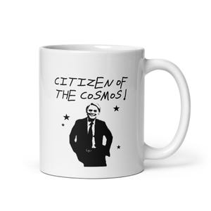 Pode incluir: Uma caneca de cerâmica branca com uma estampa gráfica preta de uma pessoa de terno. Acima da figura, o texto diz CITIZEN OF THE COSMOS! em estilo manuscrito. Várias pequenas estrelas pretas estão espalhadas pela superfície da caneca.