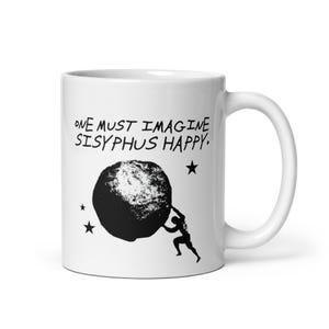 May include: A white ceramic mug featuring a black graphic illustration of a figure pushing a large boulder. Above the illustration, the text reads, "ONE MUST IMAGINE SISYPHUS HAPPY." Three small black stars are positioned around the central design.