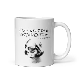 Può includere: Una tazza in ceramica bianca con un ritratto in bianco e nero di una persona con i capelli corti che guarda verso il basso. Sopra il ritratto, il testo recita: I AM A VICTIM OF INTROSPECTION. - SYLVIA PLATH. La tazza ha un manico curvo a destra.