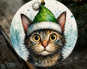 May include: A round ceramic ornament featuring a watercolor illustration of a tabby cat with large yellow eyes wearing a green and white elf hat. The background is a light blue wash with white snowflake accents and a gold hanging string.
