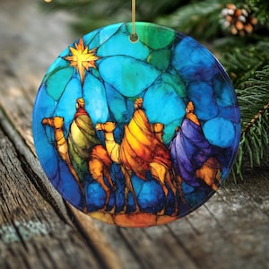 May include: Round Christmas ornament featuring a stained-glass style depiction of the Three Wise Men on camels, guided by a star. The ornament has a blue, green, and yellow color scheme, with a gold string for hanging. A festive holiday decoration.