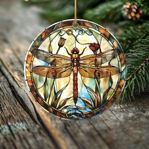 May include: Round stained glass ornament featuring a dragonfly. The dragonfly has orange and brown wings and a red head. The ornament has a gold string for hanging and is surrounded by a colorful floral design.