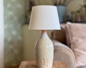 Può includere: Una lampada da tavolo in ceramica color crema con paralume bianco. La base della lampada ha un effetto smaltato a goccia. È posizionata su una superficie di legno. La lampada è progettata per uso interno e fornisce illuminazione ambientale.