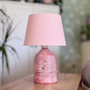 Puede incluir: Una lámpara de mesa con una base cilíndrica de cerámica con una superficie texturizada y acanalada en tonos rosa y blanco con motas oscuras. La lámpara está rematada con una pantalla de tela rosa claro lisa con una forma cónica.