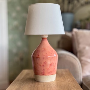 May include: A ceramic table lamp with a coral-colored base and a white lampshade. The base has a speckled, off-white bottom. The lamp is on a wooden table. The background includes a sofa and a wall with a floral pattern.