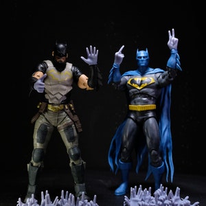Puede incluir: Dos figuras de acción de Batman, una con un traje azul y negro con un símbolo de murciélago amarillo, y la otra con un traje gris y negro con un símbolo amarillo. Ambas figuras están posadas con los brazos en alto, y hay varias pequeñas manos de plástico grises esparcidas frente a ellas.