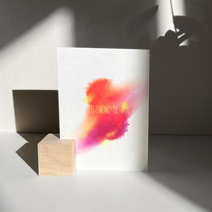 Könnte beinhalten: Eine weiße Karte mit einem Aquarelldesign in Rosa-, Orange- und Gelbtönen. Der Text "Thinking of you" ist in goldenen Buchstaben geschrieben.