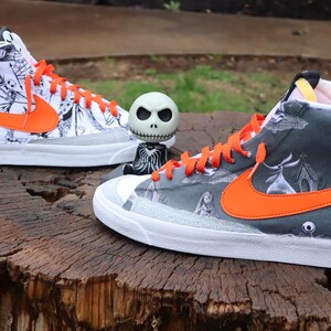 May include: A pair of high-top trainers with a white base and orange accents. The trainers feature a black and white print of characters from the film "The Nightmare Before Christmas".