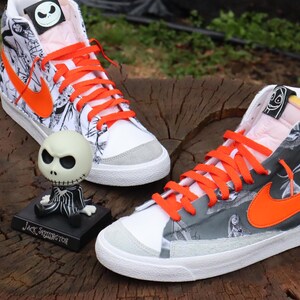 May include: A pair of white high-top trainers with black and white patterned fabric and orange laces. The trainers have a black and white Nike swoosh logo on the side. The trainers are decorated with a black and white illustration of Jack Skellington from the film The Nightmare Before Christmas. The text "Jack Skellington" is printed on the bottom of the trainers.
