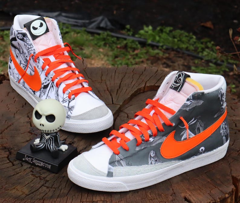 May include: A pair of white high-top trainers with black and white patterned fabric and orange laces. The trainers have a black and white Nike swoosh logo on the side. The trainers are decorated with a black and white illustration of Jack Skellington from the film The Nightmare Before Christmas. The text "Jack Skellington" is printed on the bottom of the trainers.