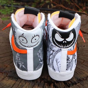 May include: A pair of white high-top trainers with a black and white print of characters from The Nightmare Before Christmas. The left shoe features Sally's face and the right shoe features Jack Skellington's face. The shoes have orange accents.