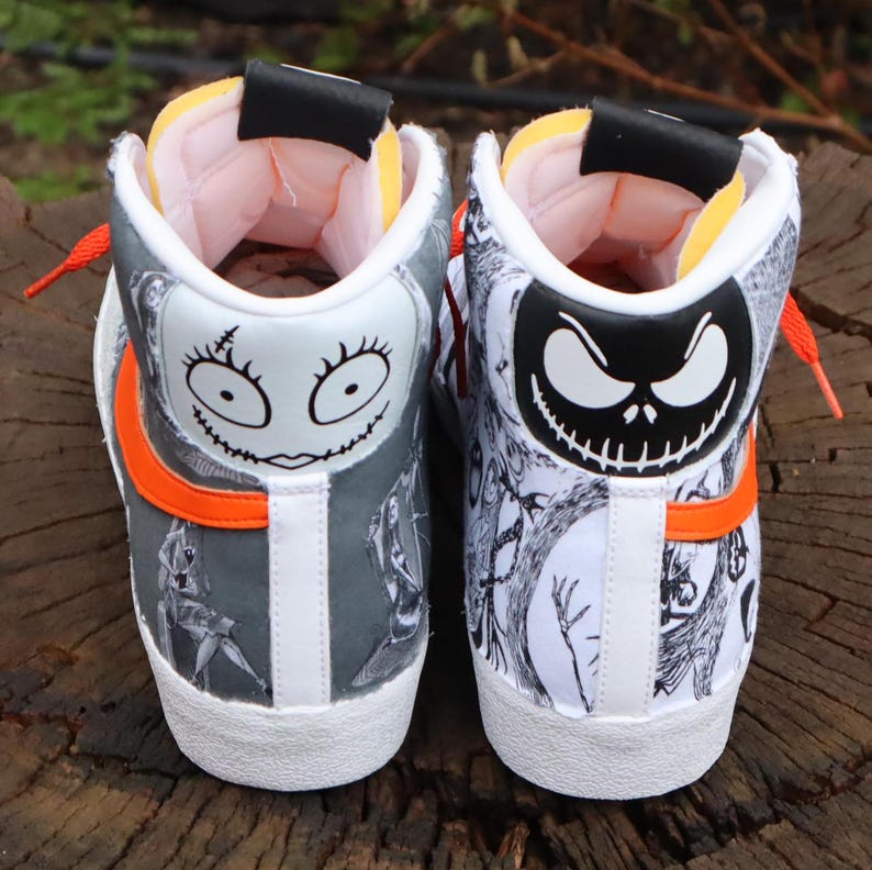 May include: A pair of white high-top trainers with a black and white print of characters from The Nightmare Before Christmas. The left shoe features Sally's face and the right shoe features Jack Skellington's face. The shoes have orange accents.
