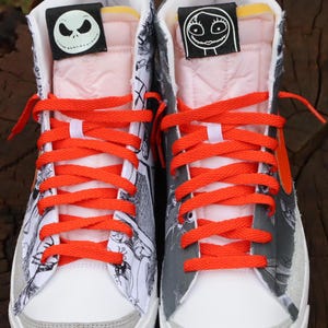 May include: A pair of white high-top trainers with black and white cartoon illustrations. The trainers have orange laces and a grey suede toe. The tongue of each shoe has a black patch with a white cartoon character. The left shoe has a patch with Jack Skellington from The Nightmare Before Christmas. The right shoe has a patch with Sally from The Nightmare Before Christmas.