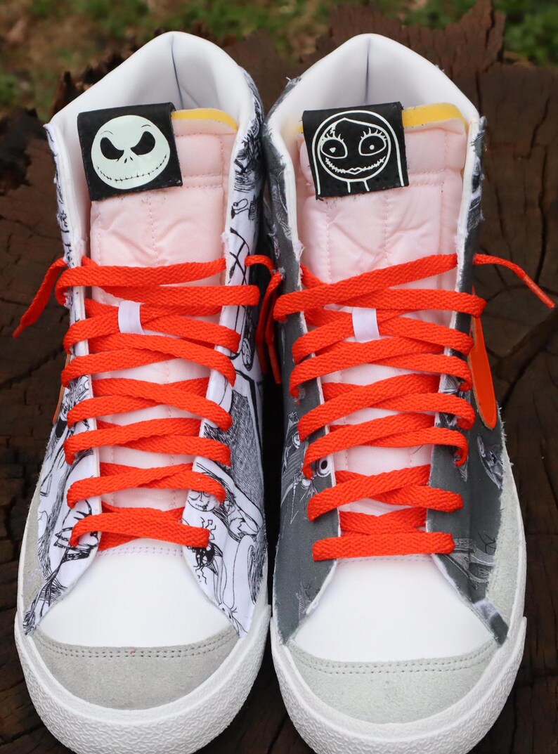 May include: A pair of white high-top trainers with black and white cartoon illustrations. The trainers have orange laces and a grey suede toe. The tongue of each shoe has a black patch with a white cartoon character. The left shoe has a patch with Jack Skellington from The Nightmare Before Christmas. The right shoe has a patch with Sally from The Nightmare Before Christmas.