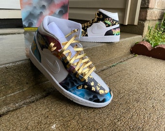 May include: A pair of white high-top sneakers with a custom paint design. The sneakers feature a blue, black, and gold paint design with gold accents. The laces are gold.