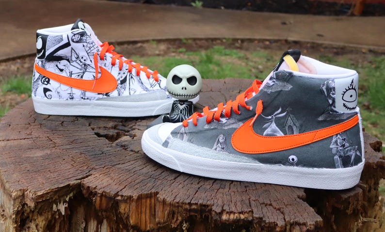 May include: A pair of high-top trainers with a white base and orange accents. The trainers feature a black and white print of characters from the film "The Nightmare Before Christmas".