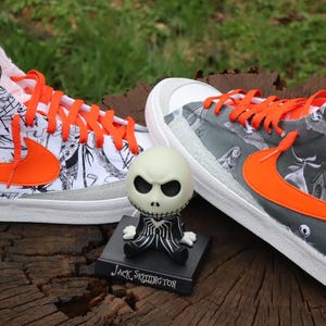 May include: A pair of white high-top trainers with black and white illustrations of characters from the film "The Nightmare Before Christmas". The trainers have orange laces and a bright orange swoosh logo. A small Jack Skellington figurine is in front of the trainers.
