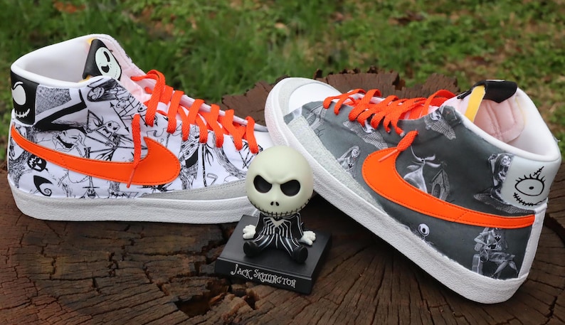 May include: A pair of white high-top trainers with black and white illustrations of characters from the film "The Nightmare Before Christmas". The trainers have orange laces and a bright orange swoosh logo. A small Jack Skellington figurine is in front of the trainers.