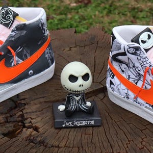 May include: A pair of high-top trainers with a white base and black and white illustrations of characters from The Nightmare Before Christmas. The trainers have orange accents, including the Nike swoosh and laces. The trainers are sitting on a wooden surface with a small Jack Skellington figurine in front of them.