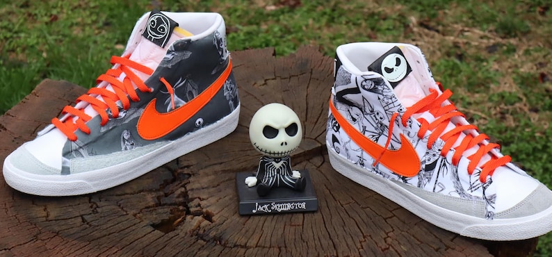 May include: A pair of high-top trainers with a white base and black and white illustrations of characters from The Nightmare Before Christmas. The trainers have orange accents, including the Nike swoosh and laces. The trainers are sitting on a wooden surface with a small Jack Skellington figurine in front of them.