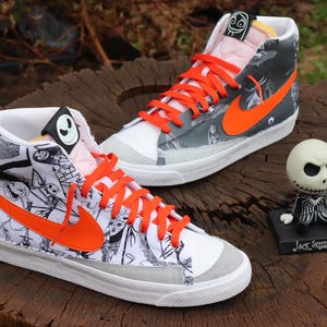May include: A pair of white high-top trainers with black and white illustrations of characters from The Nightmare Before Christmas. The trainers have orange laces and a bright orange Nike swoosh. The right trainer has a black and white illustration of Jack Skellington's face on the tongue.