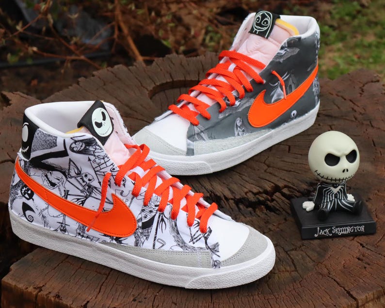 May include: A pair of white high-top trainers with black and white illustrations of characters from The Nightmare Before Christmas. The trainers have orange laces and a bright orange Nike swoosh. The right trainer has a black and white illustration of Jack Skellington's face on the tongue.