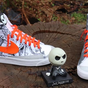 May include: A pair of white high-top trainers with orange laces and a black and white graphic print featuring characters from the film 'The Nightmare Before Christmas'. The trainers are sitting on a wooden surface with a small Jack Skellington figurine in front of them.