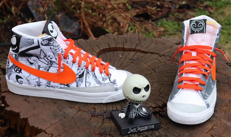 May include: A pair of white high-top trainers with orange laces and a black and white graphic print featuring characters from the film 'The Nightmare Before Christmas'. The trainers are sitting on a wooden surface with a small Jack Skellington figurine in front of them.