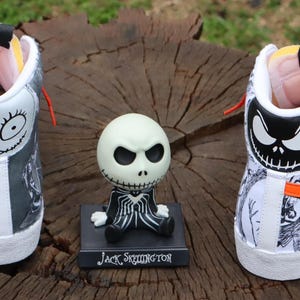 May include: A pair of white high-top trainers with orange and black accents. The trainers are decorated with a black and white illustration of Jack Skellington from The Nightmare Before Christmas. The trainers are sitting on a wooden surface next to a small Jack Skellington figurine.