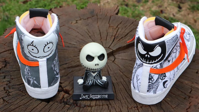 May include: A pair of white high-top trainers with orange and black accents. The trainers are decorated with a black and white illustration of Jack Skellington from The Nightmare Before Christmas. The trainers are sitting on a wooden surface next to a small Jack Skellington figurine.