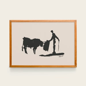 May include: Black and white artwork of a bullfight scene. A bull and a matador are depicted in a minimalist style. The artwork is framed in a light brown wooden frame.
