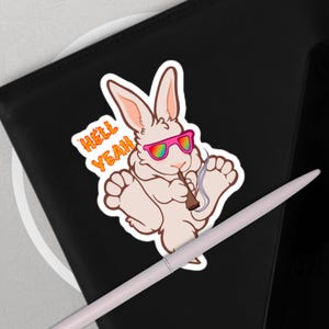 May include: A sticker featuring a cartoon rabbit with pink sunglasses and a lit cigar. The rabbit is light beige with pink ears and paws. The text "HELL YEAH" is in orange and yellow. The sticker is on a black surface.