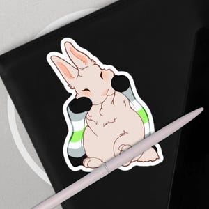 Puede incluir: Una pegatina con un conejo de dibujos animados con un cuerpo rosa claro, detalles en negro y un diseño de bandera del orgullo aroace. La pegatina está sobre una superficie negra con un bolígrafo blanco.