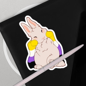 Puede incluir: Una pegatina con un conejo de dibujos animados con una bandera del orgullo no binario. El conejo es de color rosa claro con detalles en amarillo y morado. La pegatina está sobre una superficie negra con un lápiz óptico blanco.