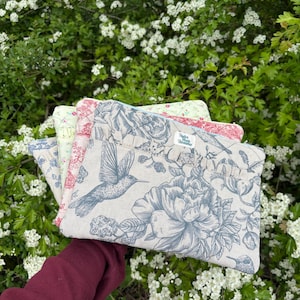 Può includere: Un gruppo di quattro pochette in tessuto con disegni floreali e colibrì. Le pochette presentano una varietà di colori, tra cui blu, rosso e verde, con uno sfondo color crema. Le pochette sono tenute davanti a uno sfondo di fiori bianchi e fogliame verde.