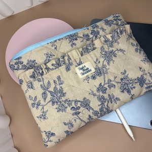May include: Beige quilted tablet sleeve with a blue floral pattern and a light blue zipper. The sleeve has a tag that reads "Taylor Made Treasures". A tablet and stylus are visible.
