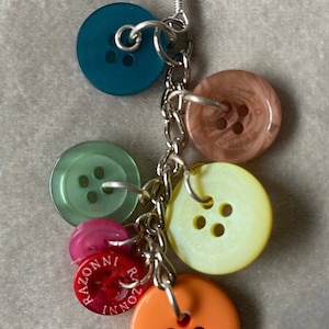 May include: A silver chain earring with a variety of colourful buttons attached. The buttons are in shades of teal, green, brown, yellow, pink, and orange. Each button has four holes and a silver loop attached.