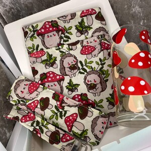 May include: A set of three items featuring a hedgehog and mushroom pattern. The set includes a fabric pouch, a fabric wristlet, and a glass cup. The pouch and wristlet are both cream coloured with a repeating pattern of hedgehogs, mushrooms, and leaves. The glass cup is clear with a red and white mushroom design.