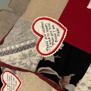 Pode incluir: Uma almofada de patchwork com v&aacute;rios padr&otilde;es e cores de tecido, incluindo vermelho, preto floral e cinza. Um patch em forma de cora&ccedil;&atilde;o com texto bordado diz: "Estas s&atilde;o as roupas que eu usava quando voc&ecirc; me abra&ccedil;a, sei que estou perto."