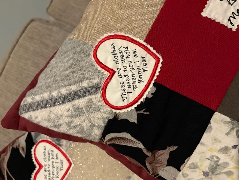 Pode incluir: Uma almofada de patchwork com v&aacute;rios padr&otilde;es e cores de tecido, incluindo vermelho, preto floral e cinza. Um patch em forma de cora&ccedil;&atilde;o com texto bordado diz: "Estas s&atilde;o as roupas que eu usava quando voc&ecirc; me abra&ccedil;a, sei que estou perto."