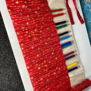 Puede incluir: Un rollo de lápices de tela roja con estrellas doradas y un forro blanco. El rollo está desenrollado y muestra una variedad de lápices de colores en el interior.