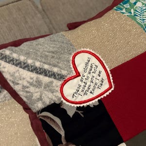 Pode incluir: Uma almofada de patchwork com quadrados de tecido em tons de cinza, vermelho, preto e verde. Um patch em forma de cora&ccedil;&atilde;o com o texto bordado: "These are clothes I used to wear when you hold Know I am Near."