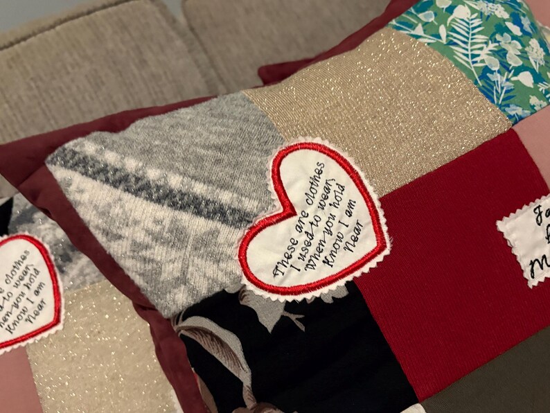 Pode incluir: Uma almofada de patchwork com quadrados de tecido em tons de cinza, vermelho, preto e verde. Um patch em forma de cora&ccedil;&atilde;o com o texto bordado: "These are clothes I used to wear when you hold Know I am Near."