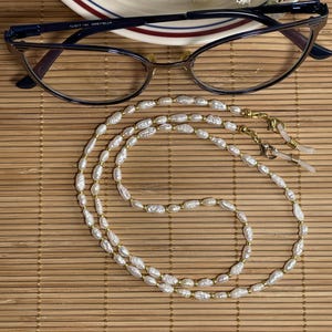 May include: A pair of navy blue eyeglasses with a gold-colored nose bridge and a white pearl eyeglass chain. The chain is made of white pearls and gold-colored beads. The eyeglasses are on a bamboo mat.