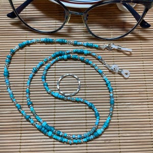 May include: A pair of eyeglasses with dark blue frames rests above a beaded eyeglass chain. The chain is turquoise and silver, with small beads and silver clasps. A silver ring is also present.
