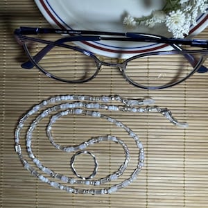 May include: A white and silver beaded eyeglass chain with a silver ring. The chain is made of small, round beads and rectangular beads. The chain is lying on a brown woven surface.