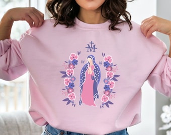 May include: A pink sweatshirt with a floral design and a depiction of the Virgin Mary. The design includes a cross and the letters "M" and "A" in a stylized font.