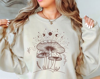 May include: A cream-colored sweatshirt with a brown mushroom graphic and celestial symbols. The sweatshirt has long sleeves and a relaxed fit.